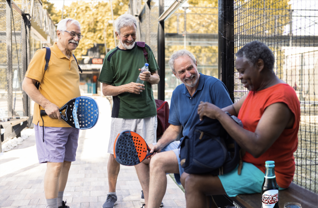 A group of diverse people laughing and playing pickleball with one person munching on a snack